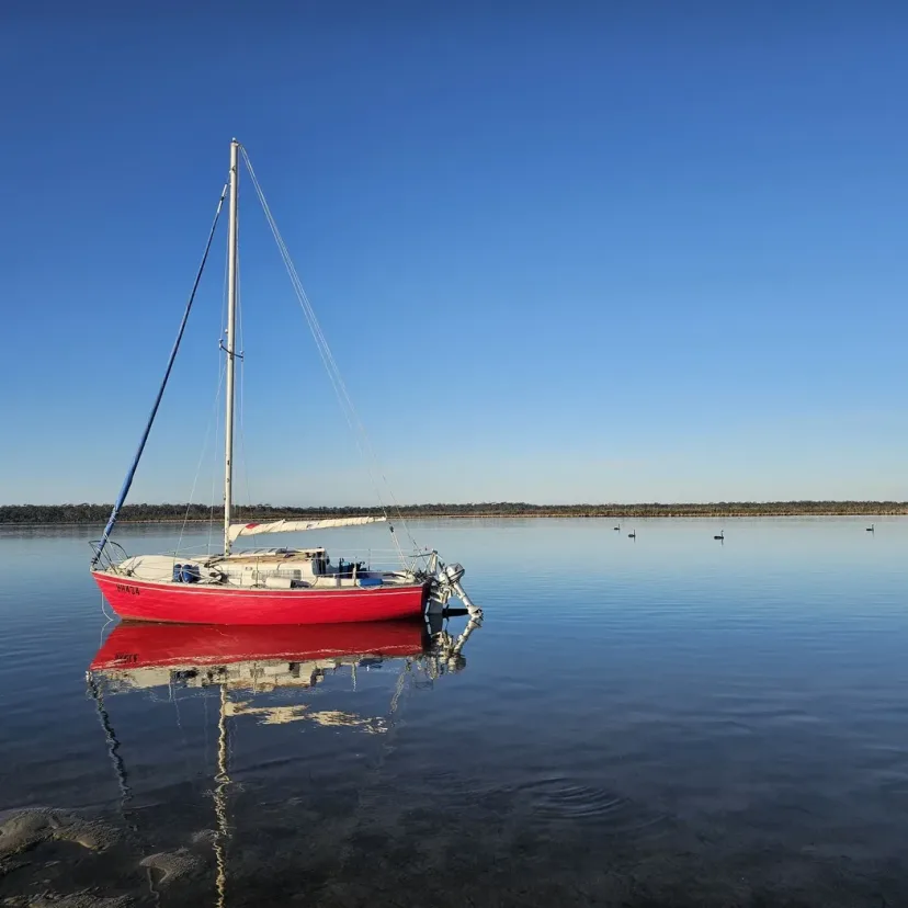 A red hulled yacht moored on flat calm water near a wooded shore